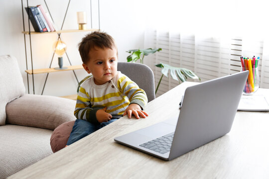 Two Year Old Boy Staring At The Laptop Screen With Fascinated Facial Expression. Toddler Boy Sitting By The Table At Home With Notebook Computer. Close Up, Copy Space, Background.