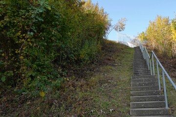 Fototapeta premium Emergency staircase from an emergency exit on a highway. The staircase is made of cement and has a metal railings on one side, it is bordered on both sides by bushes and trees in autumn colors.