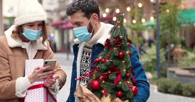 Close up portrait of Caucasian happy couple in masks in decorated city with christmas presents taking selfie photo on smartphone while snowing. Joyful wife and husband outdoor on Christmas eve