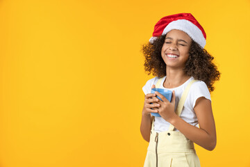 Happy little girl in Santa hat laughing with cup of hot drink isolated on yellow background