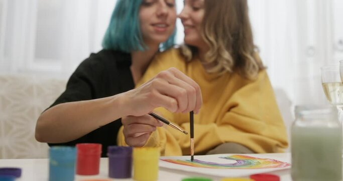 Loving lesbian couple painting together at home. Young lesbian women kissing and painting rainbow on paper while sitting at table with paintbrushes and spending time together