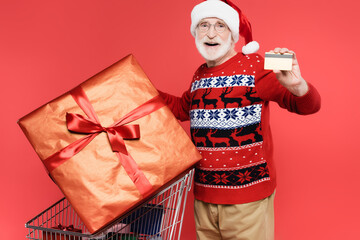 Fototapeta premium Smiling elderly man in santa hat holding credit card near gifts in shopping cart on red background
