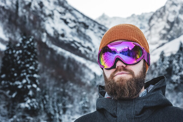 Fototapeta na wymiar An adult brutal man with a beard in ski goggles and a hat stands in the mountains in winter. Active recreation concept