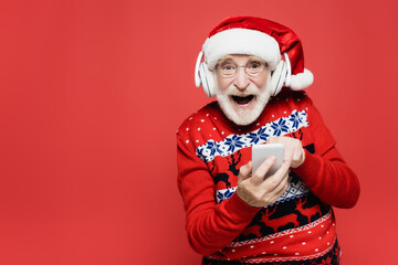Cheerful senior man in santa hat using smartphone and wireless headphones isolated on red
