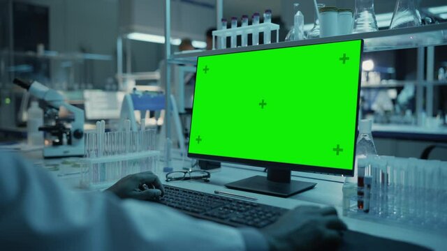 Medical Research Scientist Working on Desktop Computer with Green Screen Mock Up Template in Applied Science Research Laboratory. Lab Engineers in White Coats Conduct Experiments in the Background.