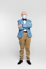 Senior man in medical mask standing with crossed arms on grey background