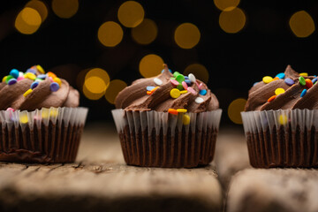 Tasty cupcakes with chocolate cream and colored sprinkles on rustic wooden table on black background. Sweet dessert. Bakery concept. Elegant food. Sweets for coffee or tea.