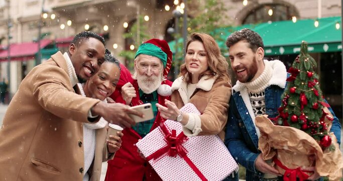 Portrait of old Santa Claus hugging happy African American and Caucasian females and males while taking selfie photo on smartphone. Mixed-races joyful friends with Santa Clause on street. X-mas mood