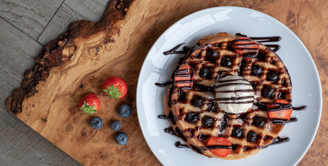 Vegan waffle with vanilla icecream, chocolate sauce, and decorated with blueberries and strawberries. Photographed from above on an olive wood platter.