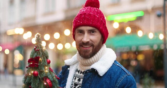 Close up portrait of cheerful Caucasian man in red warm hat standing in decorated town with small new year's tree in hands. Portrait of handsome male on street on xmas day. Winter concept