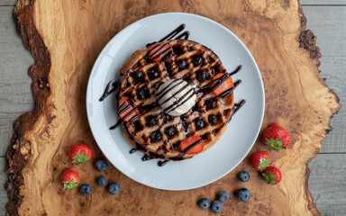 Vegan waffle with vanilla icecream, chocolate sauce, and decorated with blueberries and strawberries. Photographed from above on an olive wood platter.