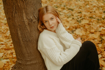 A girl in a white sweater sits on the autumn leaves. A beautiful blonde with a square looks at the camera. Woman outdoors in the autumn park.