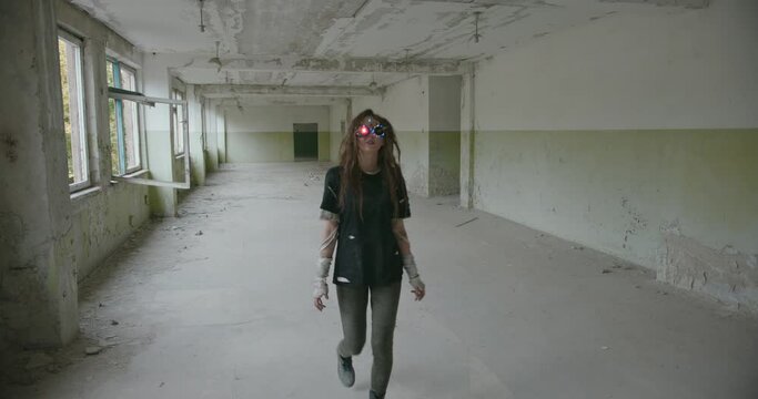Woman in dirty clothes and VR glasses in abandoned building. Mad female inventor with messy hair wearing handmade grunge VR goggles walking towards camera and smiling