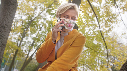 Young fashional woman with fabric face mask having phone conversation in the park under the tree on autumn day. High quality photo
