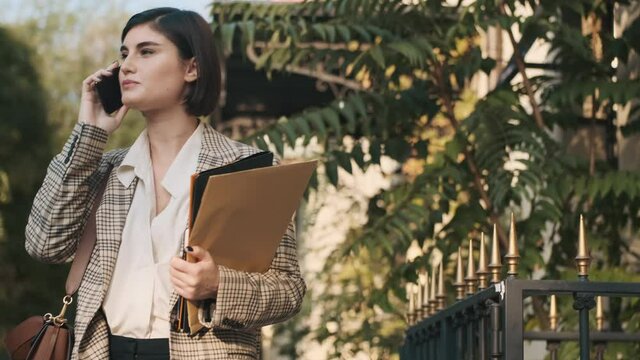 Young beautiful female architector dressed in plaid blazer talking on smartphone while waiting customer on city street. Businesswoman working outdoor