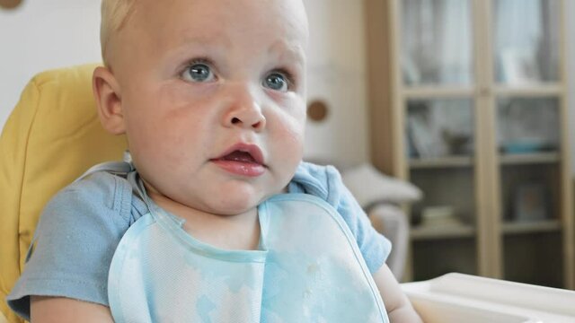 Close up of adorable child with blond hair and blue eyes wearing bib sitting at highchair at home and looking around