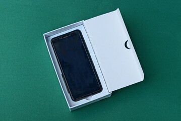 one black smartphone lies in a white open paper box packaging on a green table
