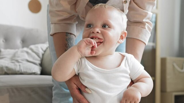 Close up of unrecognizable young mother wearing casual clothes standing in living room and holding adorable cheerful little son trying to stand straight