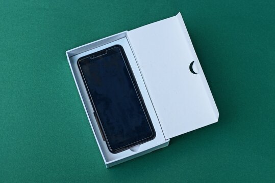 one black smartphone lies in a white open paper box packaging on a green table