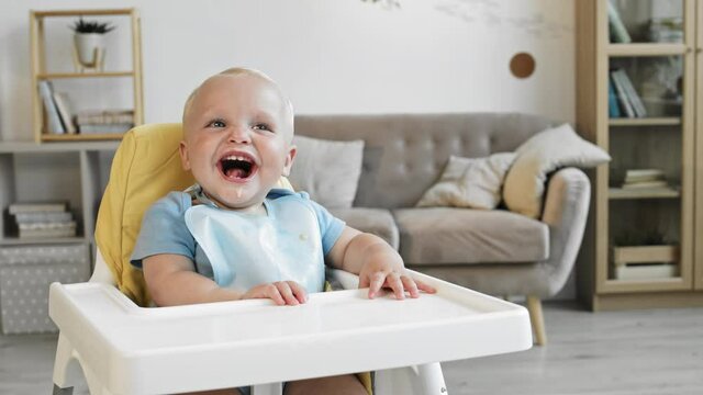 Medium shot of adorable baby boy with blond hair and blue eyes wearing burp cloth sitting at highchair at home and laughing