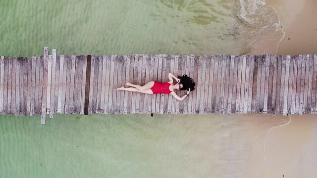 aerial view of asian woman in bikini lay down on wooden bridge over the transparent sea water near beach. copy space provided. travel concept. Thailand.