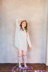 Naklejka premium Girl in a long light sweater, jacket, and garland with Christmas lights, Scandinavian style, new year, waiting, fun, holiday, friends