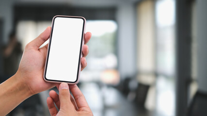 Cropped shot hands holding blank screen smartphone with blurred interior of modern office background.