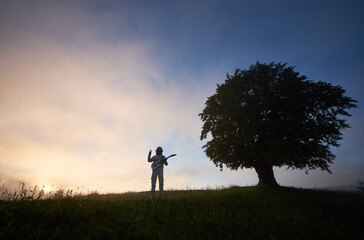 Two figures on purple sky background, of man astronaut in space suit holding guitar and showing peace sign and a big old tree on the right © anatoliy_gleb