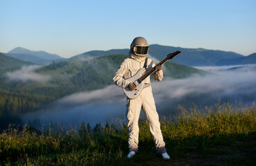 Naklejka premium Astronaut in white space suit and helmet playing guitar, standing on sunny green mountain glade in summer, morning fog rising up from the valley behind him. Concept of astronautics, music and nature.