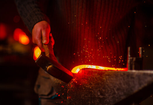 The blacksmith hits the red-hot workpiece in the forge with a hammer and glowing sparks fly in all directions