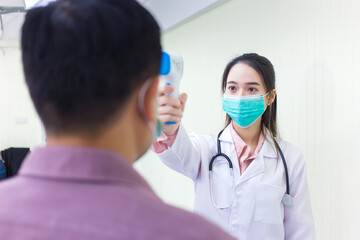 Asian woman doctor uses a infrared forehead thermometer to check body temperature of a man patient in hospital. Both of them wear medical face mask to protect pathogen in health care concept.