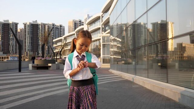 Little child girl with a backpack in school uniform is going to school in city landscape and checking the time on watch on her hand, front view. Primary school pupil is going to study, steadicam shot.
