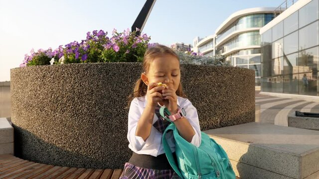 Little child girl with a backpack in school uniform is sitting on bench and eating the apple in school way.