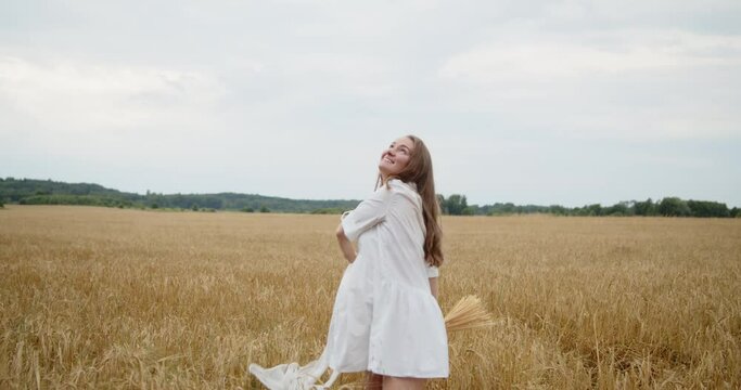 Young pregnant woman in a white summer dress spinning around and waving hands in a wheat field holding bunch of wheat and hat. Slow motion wide shot.