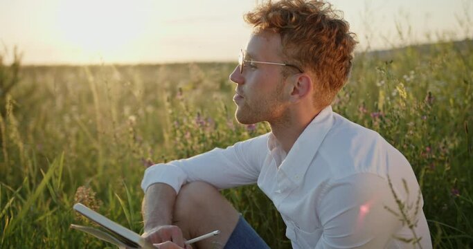 Guy writer artist dressed in a shirt writes himself with inspiration with pleasure writes poems and poetry in nature in a field of flowers outside the city at sunset inspired by the beauty of nature