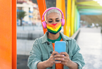 Young woman wearing gay pride mask using mobile smartphone while listening to music with wireless headphones outdoor - Gender equality and technology concept