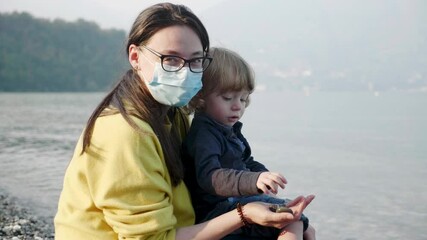 Millennial mother with face covering plays with her cute toddler son at the edge of Lake Garda - Powered by Adobe