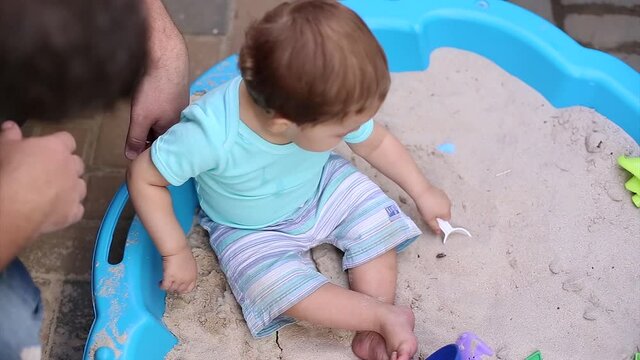 Little baby boy sits on sand in sandbox and plays with colorful toys on sandy background