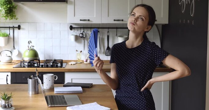 Overheated young stressed woman standing in kitchen, using paper fan, suffering from extremely hot summer weather without conditioning system at home, cooling herself, breathing fresh air indoors.