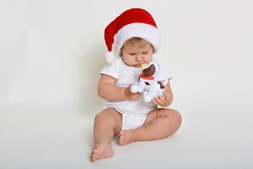 Cute baby wearing christmas hat and white bodysuit playing with plastic toy dog while sitting barefoot on floor, looks at his favorite toy with concentrated facial expression.