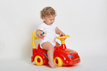 Happy smiling blond infant with curly hair driving toy car indoor against white wall, looking at rudder, wearing bodysuit, having fun with his toys, looks excited and satisfied.