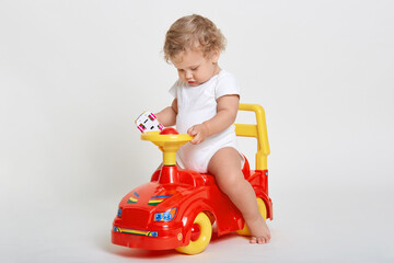 Little charming baby boy sitting on red and yellow tolocar, holding toy car in hands, wearing white body suit, has blond curly hair, cute infant playing indoor.