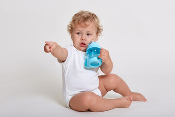 Little boy drinking water while sitting on floor isolated over white background, looking and pointing away, wearing body suit, has blond curly hair, child with blue baby cup.