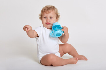 Charming baby wants to drink water from bottle, looking away, pointing with his index finger, wearing white body suit, sitting on floor against white wall.
