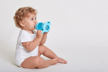 Portrait of cute toddler drinking water from bottle while sitting against white wall, wearing body suit, one year old kid with curly hair being thirsty, side view of toddler.