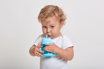 Little boy drinking water from bottle, wearing t shirt, looking directly at camera, posing against white background, male child with blond curly hair.