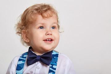 Cute smiling blonde baby kid boy in white shirt with bow tie and suspenders, looking aside over white background, having curious expression, copy space for advertisement.