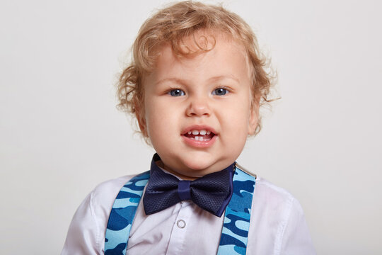 Little Handsome Curly Boy With Big Beautiful In White Shirt With Bow Tie Stands In Room And Blue Suspenders, Looks At Camera, Shows His Teeth, Posing Isolated Over White Background.