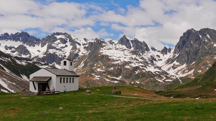 Fototapeta na wymiar Hikers resting on benches in front of a small chapel near Silvretta reservoir, Montafon, Austria with beautiful mountain panorama in background with snow-capped mountains in early summer.