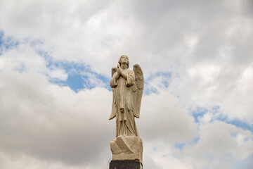 Escultura de anjo em topo de mausoléu dentro de um cemitério com céu nublado ao fundo.